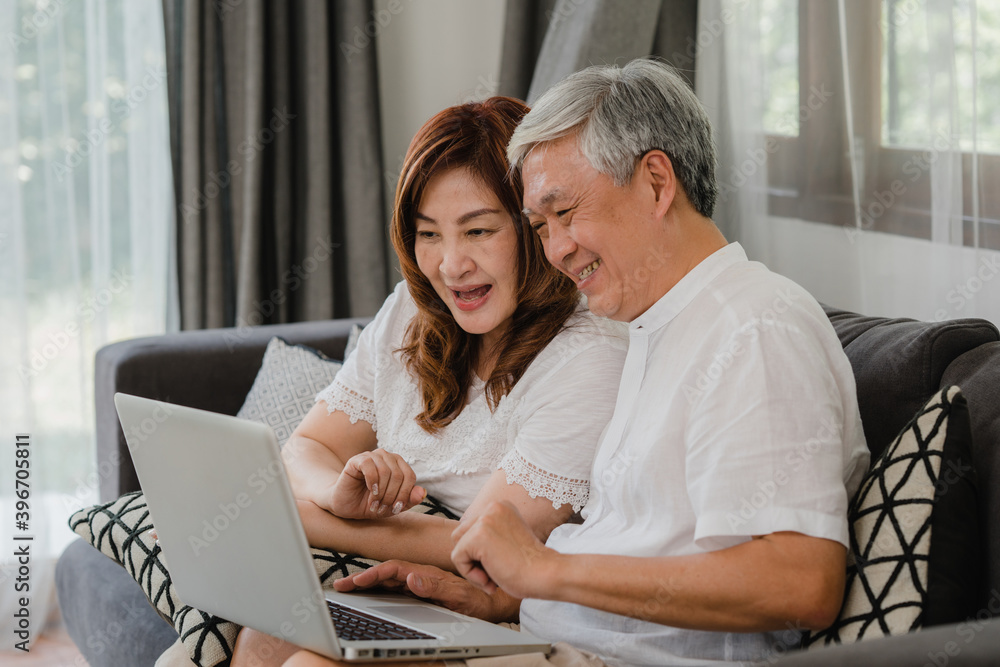 Asian senior couple using laptop at home. Asian Senior Chinese grandparents, surf the Internet to check social media while lying on sofa in living room at home concept.