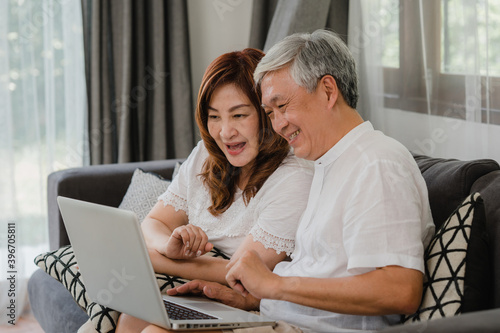 Asian senior couple using laptop at home. Asian Senior Chinese grandparents, surf the Internet to check social media while lying on sofa in living room at home concept.