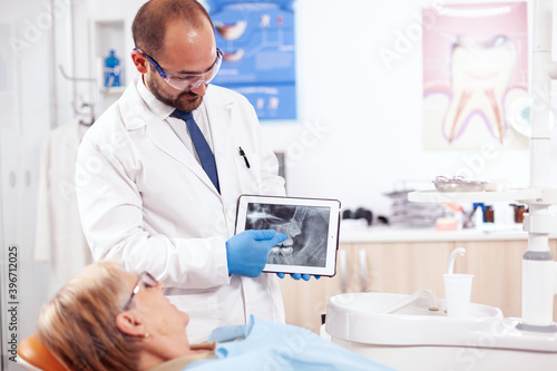 Dentist in dental cabinet explaining tooth diagnosis on digital device standing up. Medical teeth care taker holding patient radiography on tablet pc near patient standing up.