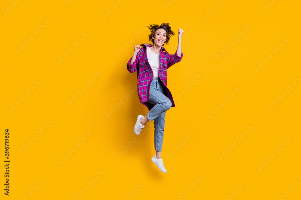 Full size photo of triumphant happy girl wear jeans violet checkered cardigan raise fists jump air isolated on yellow color background Full size photo of triumphant happy girl wear jeans violet checkered cardigan raise fists jump air isolated on yellow color background