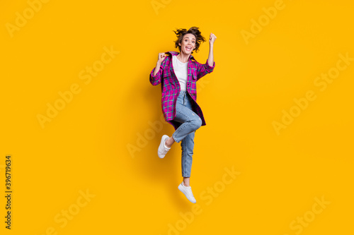 Fototapeta Naklejka Na Ścianę i Meble -  Full size photo of triumphant happy girl wear jeans violet checkered cardigan raise fists jump air isolated on yellow color background