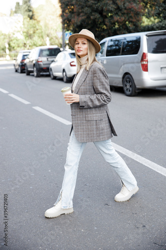 A Woman in a grey jacket and hat crosses the road with cars. A blonde girl holds a glass of coffee. Street style