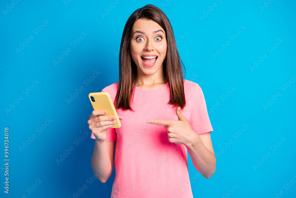 Photo portrait of female blogger pointing finger at mobile phone staring isolated on bright blue color background Photo portrait of female blogger pointing finger at mobile phone staring isolated on bright blue color background