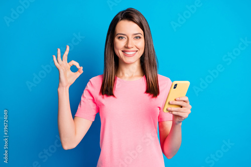 Photo portrait of female blogger using smartphone showing okay gesture smiling isolated on vivid blue color background © deagreez