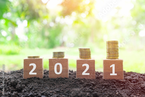 Year 2021 economy growth and recovery, increase money saving and investment concept. Wooden blocks 2021 with increasing stack of coins at sunrise on natural background.