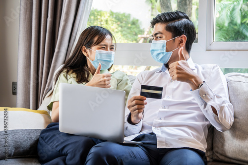 Quarantine Shopping. Young couple wearing protective masks purchasing goods online in internet with credit card and computer laptop showing thumbs up gesture together at home