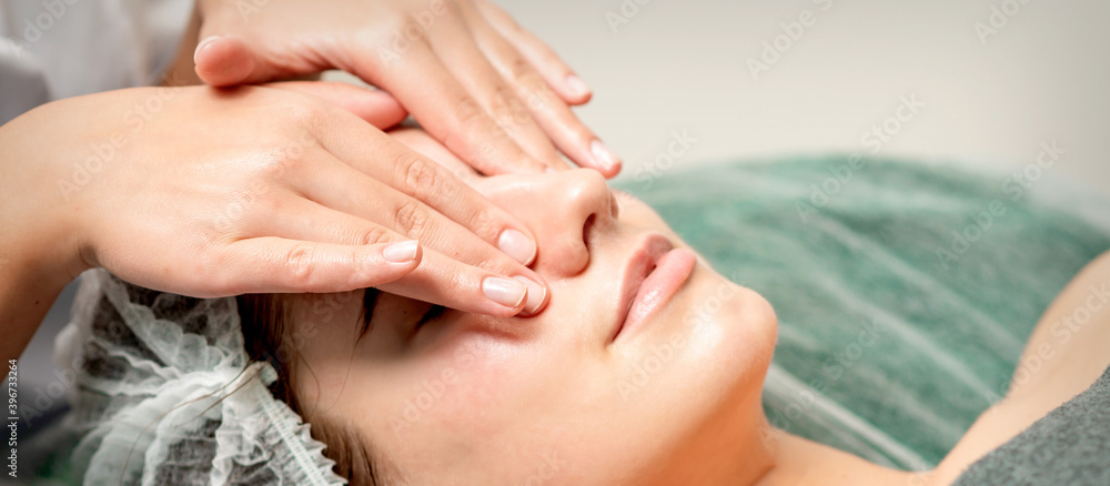 Naklejka premium Young woman getting a facial massage with closed eyes by a beautician in a beauty salon