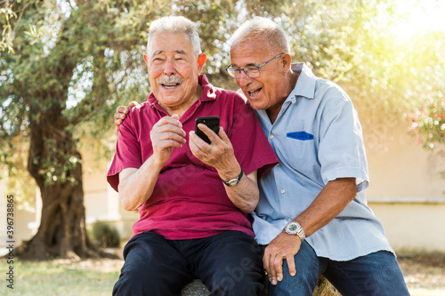Due vecchi amici scherzano e ridono davanti a uno smartphone seduti in un parco natura © alex.pin