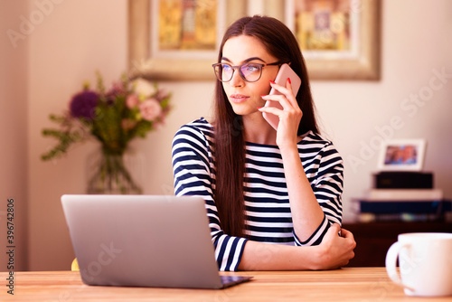 Smiling woman talking with somebody on mobile phone while sitting behind her laptop in home office