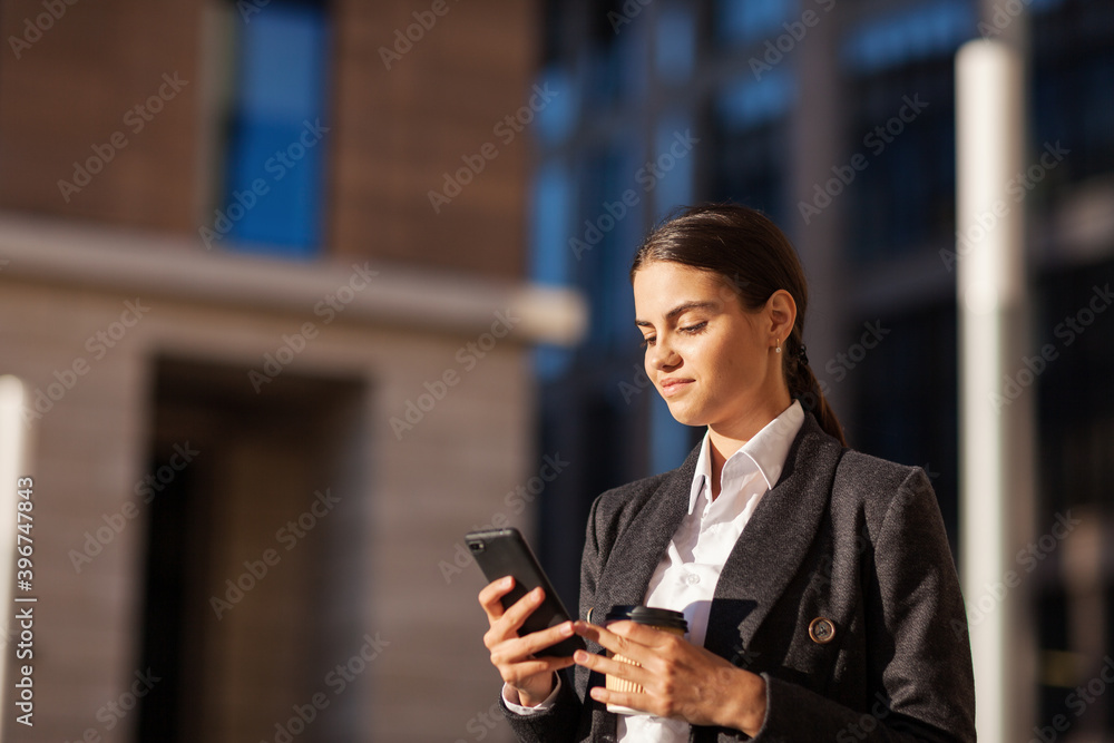 Side view of brunette young woman in suit text messaging on cell phone standing outdoors with takeaway coffee cup in her hand, copy space to left
