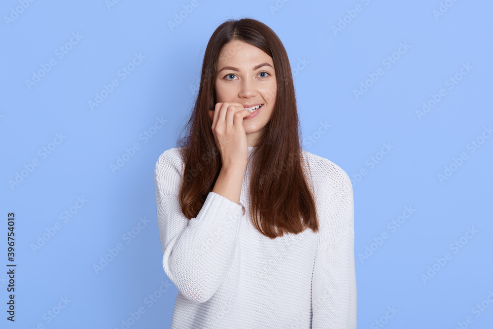 Portrait of beautiful woman biting her fingers and looking directly at camera, looks happy, wearing white casual shirt, standing against blue wall, having dark hair. Portrait of beautiful woman biting her fingers and looking directly at camera, looks happy, wearing white casual shirt, standing against blue wall, having dark hair.