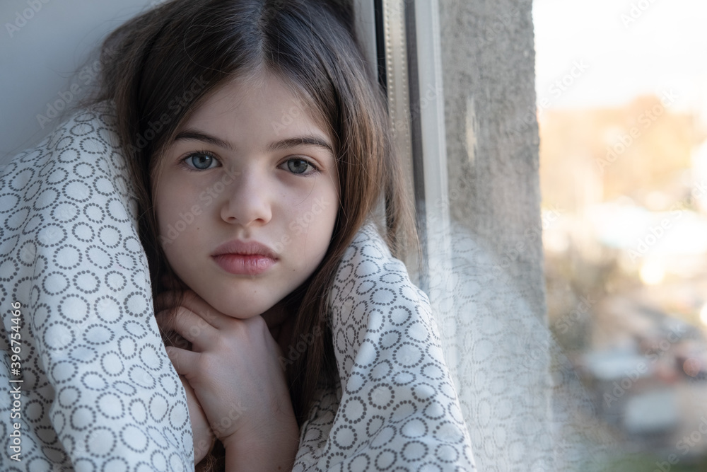 a small, young lonely sick girl in a blanket sits and looks out the window