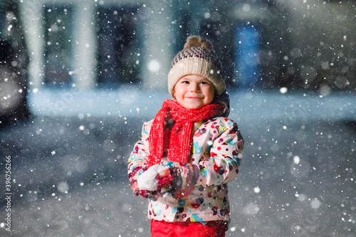 Adorable little toddler girl walking outdoors in winter. Cute toddler during strong snowfall on evening. Child having fun with snow. Wearing warm baby colorful clothes and hat with bobbles.