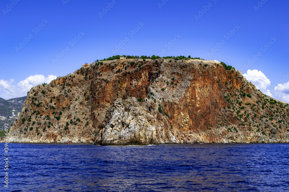 Rock with an ancient stone wall of Alanya Castle (Turkey) on top - view from the Mediterranean Sea. Seascape with an ancient fortress over a steep desert cliff against a background of blue water