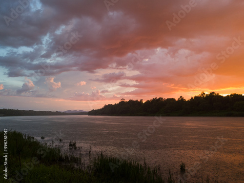 Landscape with river, colorful clouds, water grass and mountain in distance at sunset. Dramatic riverine scene during cloudy dusk with rain or summer shower and majestic beautiful glow in clouds