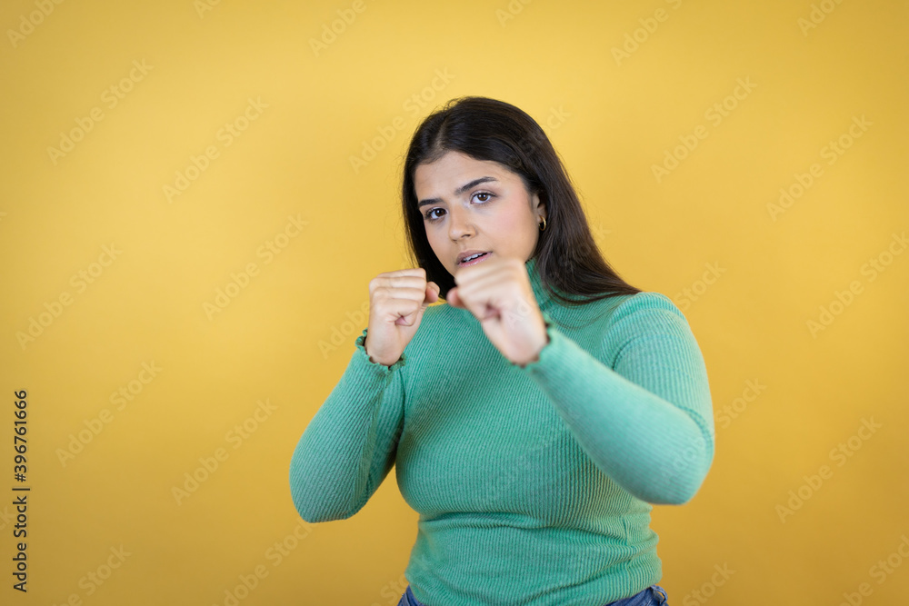 Young caucasian woman over isolated yellow background Punching fist to fight, aggressive and angry attack, threat and violence Young caucasian woman over isolated yellow background Punching fist to fight, aggressive and angry attack, threat and violence