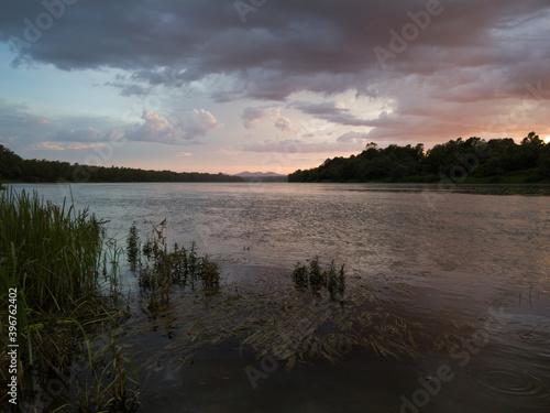Storm clouds with rain, summer shower. Landscape with river, dark clouds, tall water grass and mountain in distance at sunset. Dramatic riverine scene during cloudy dusk.