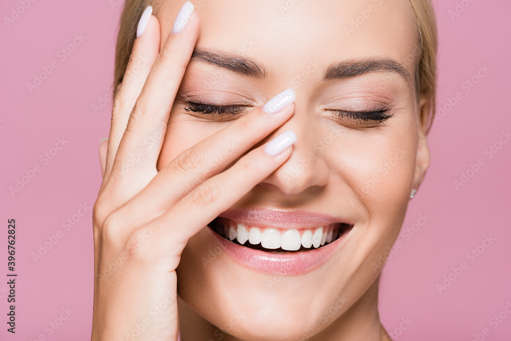 Fototapeta premium smiling beautiful blonde woman posing with hand on face isolated on pink