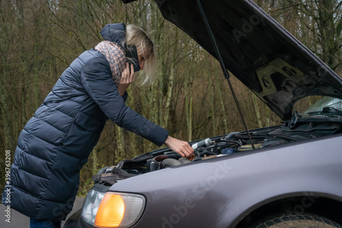 Young blonde woman in a winter down jacket in a yellow vest, talking on the phone and checking the engine oil level of the car