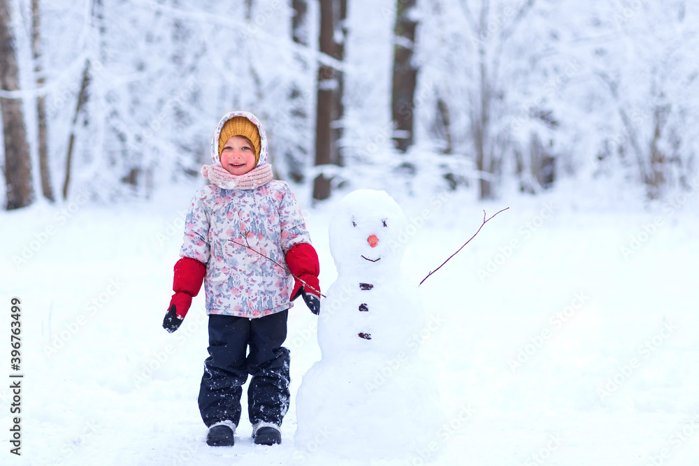 Happy child plays with a snowman on a winter walk in nature in the forest. Funny happy little girl on a walk in the winter outdoors