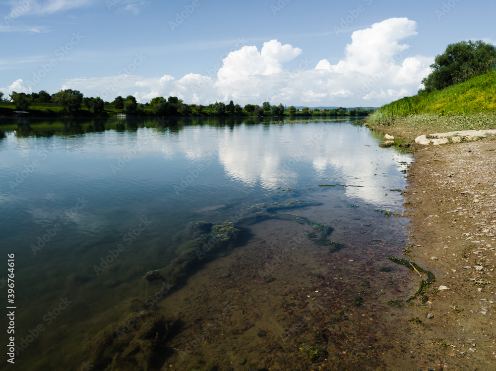 White bright cumulus clouds on blue sky. Landscape with river, grassy river bank and underwater freshwater algae White bright cumulus clouds on blue sky. Landscape with river, grassy river bank and underwater freshwater algae