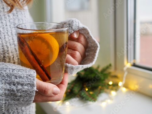 A woman's hands in a sweater hold a Cup of tea with orange and cinnamon by the window.Hot winter flavored drink. Green branches of the Christmas tree, lights