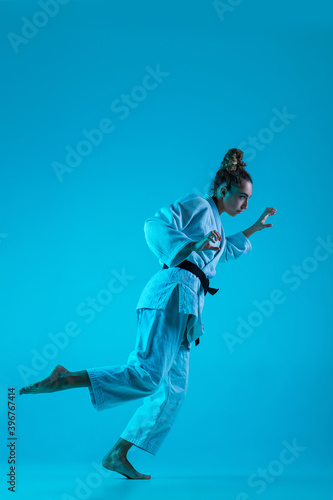 Active training. Professional female judoist in white judo kimono practicing and training isolated on blue neoned studio background. Grace of motion and action. Healthy lifestyle, sport concept