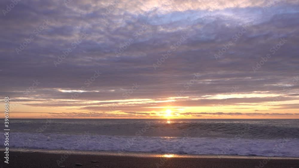 Sunset over the sea. The sun is low. The clouds are highlighted in pink, with a golden glow above the horizon. The waves roll onto the beach. White foam spreads over the sand, approaches the camera. Sunset over the sea. The sun is low. The clouds are highlighted in pink, with a golden glow above the horizon. The waves roll onto the beach. White foam spreads over the sand, approaches the camera.