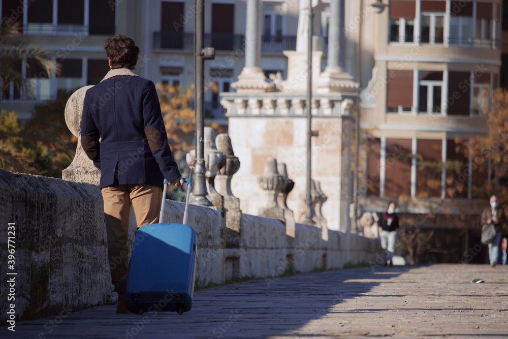 An unrecognizable young man travels to Spain during a pandemic with a blue suitcase while walking through the center of a city for work purposes. Business journey concept. Businessman 2020.