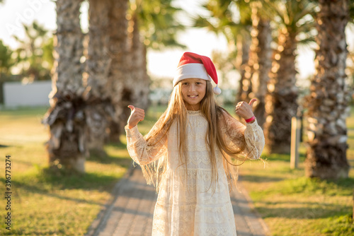 Beautiful little girl with long hair wearing a Santa Claus hat. Girl in Christmas near the palms