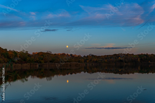 Reflection of a yellow moon on the lake during evening time. Green and orange trees surrounding the lake reflecting fall colors as well. Drak blue sky with clouds.