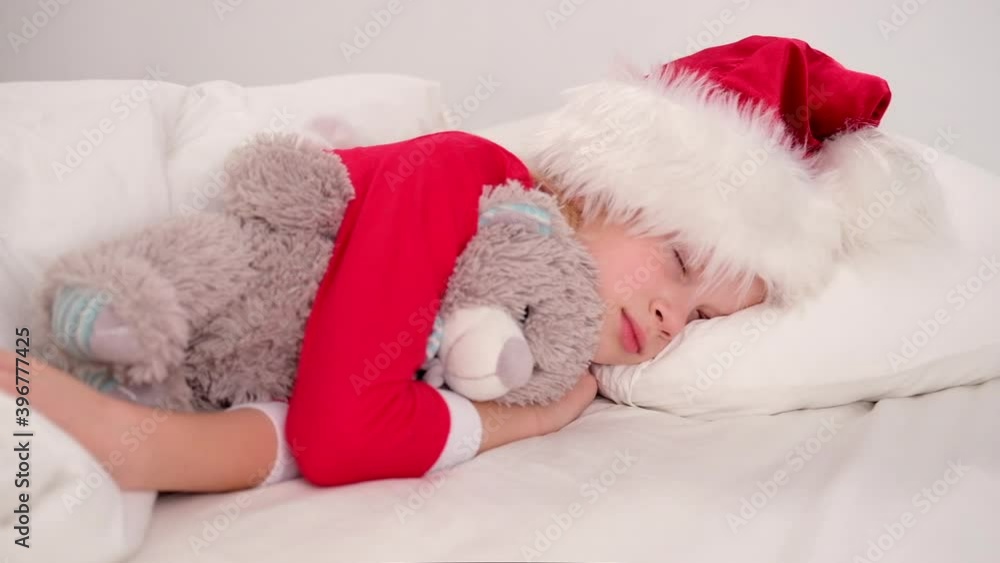 Child sleeping before Christmas. Little girl in Santa hat and Christmas pajamas lying in white bed hugging teddy bear at home. New Year and Holidays. morning happiness Child sleeping before Christmas. Little girl in Santa hat and Christmas pajamas lying in white bed hugging teddy bear at home. New Year and Holidays. morning happiness