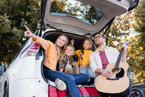 Smiling woman pointing away near children and husband with acoustic guitar in trunk of car