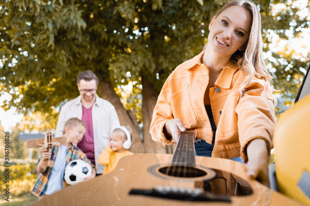 Smiling woman taking acoustic guitar from car trunk near husband and kids on blurred background outdoors