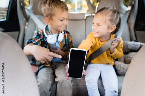 Smartphone with blank screen in hand of smiling girl and boy pointing with finger in auto on blurred background