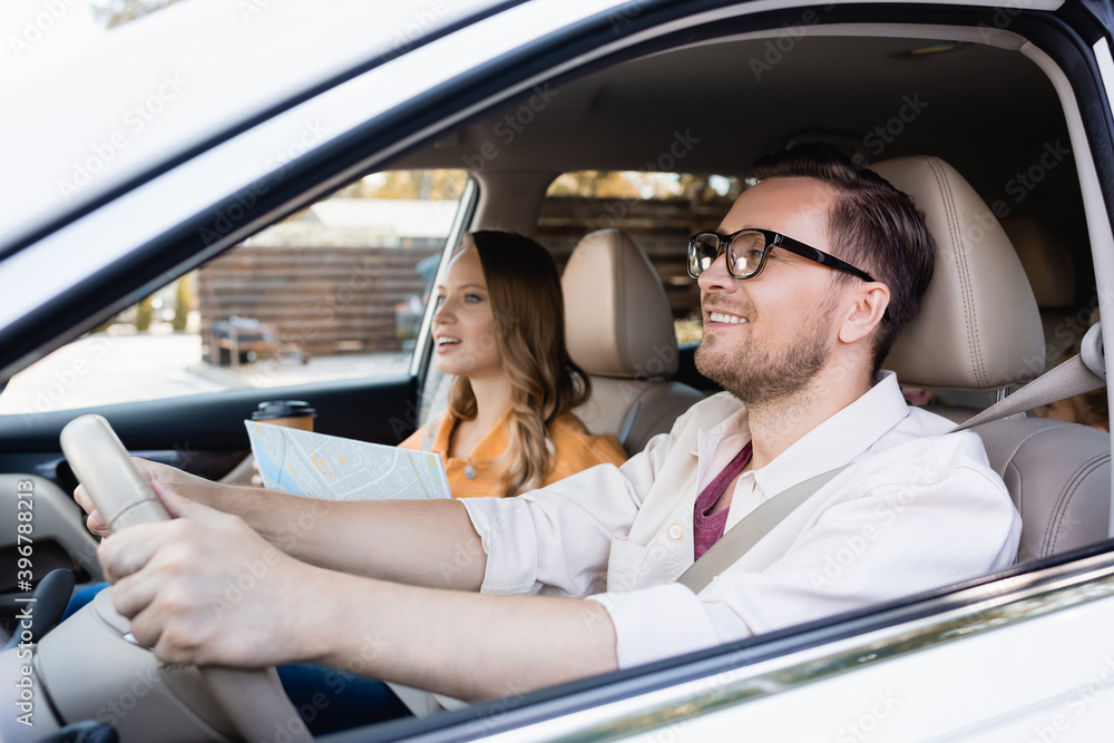 Smiling man driving car near wife with map and coffee to go on blurred background