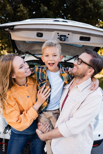 Happy kid looking at camera while hugging parents near car on blurred background outdoors © LIGHTFIELD STUDIOS
