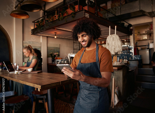Male waiter with curly hair working in cafe wearing apron with using digital tablet with female working in background on laptop photo