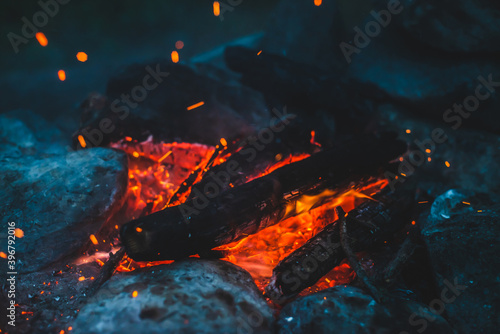 Vivid smoldered firewoods burned in fire closeup. Atmospheric background with orange flame of campfire. Wonderful full frame image of bonfire with glowing embers in air. Warm logs, bright sparks bokeh