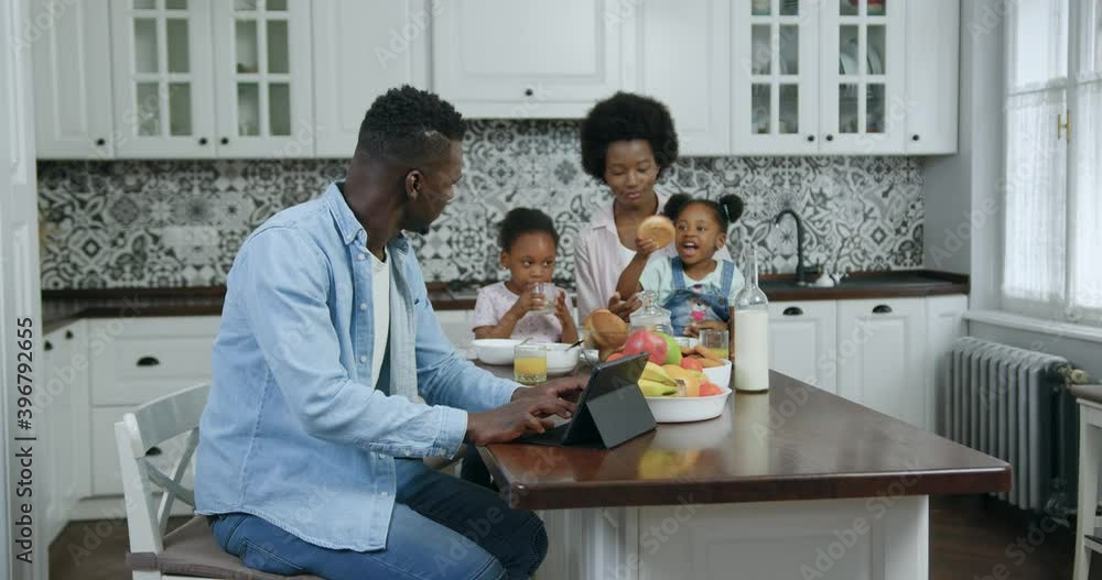 Handsome loving satisfied black-skinned man working on tablet computer at dinner table in modern kitchen while his pretty happy wife holding on knees their two kids which have breakfast,slow motion Handsome loving satisfied black-skinned man working on tablet computer at dinner table in modern kitchen while his pretty happy wife holding on knees their two kids which have breakfast,slow motion