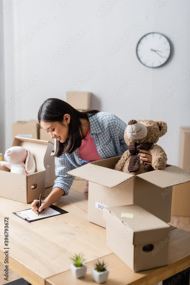 Fototapeta premium Smiling asian volunteer writing on clipboard near packages with soft toys in charity center