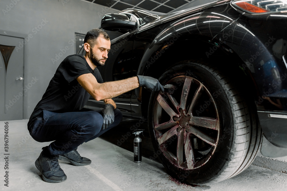 Manual wash at car wash maintance service. Handsome young male employee worker cleaning the wheel rim of modern black crossover car by scrubbing brush Manual wash at car wash maintance service. Handsome young male employee worker cleaning the wheel rim of modern black crossover car by scrubbing brush