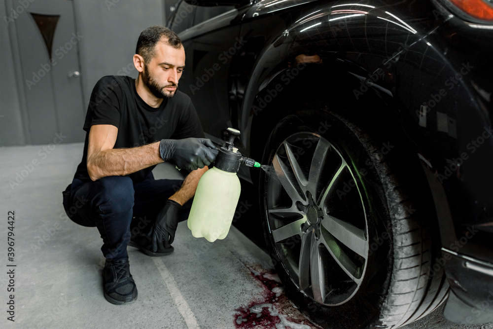 Washing a car in detailing service. Indoor shot of male worker in uniform, washing the car wheels rims with pressure water sprayer after cleaning it with special red cleansing solution