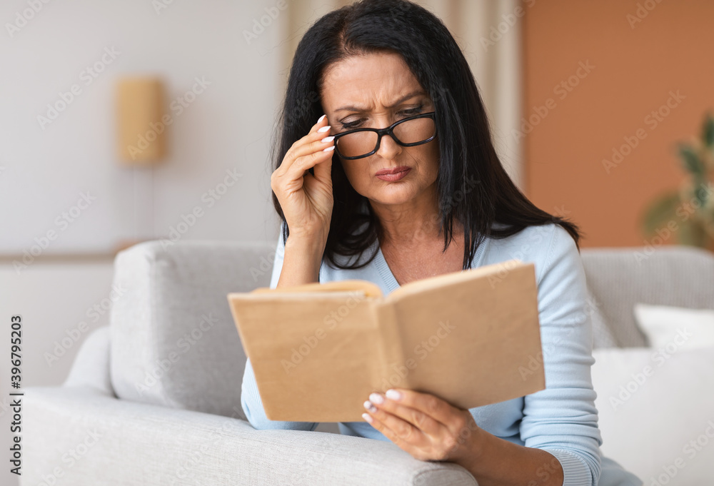 Mature woman touching glasses trying to read book Mature woman touching glasses trying to read book