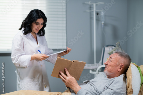 A female doctor interacting with her senior patient. The old man is showing her the book he is reading, while she is making some notes beside his bed in a hospital ward.