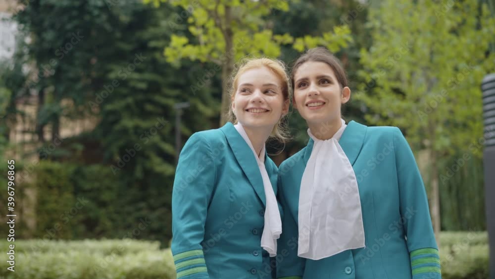 Two young beautiful women in stewardess uniform gossiping outdoors and pointing away. Portrait of slim charming Caucasian flight attendants chatting on city street. Tourism and femininity concept. Two young beautiful women in stewardess uniform gossiping outdoors and pointing away. Portrait of slim charming Caucasian flight attendants chatting on city street. Tourism and femininity concept.