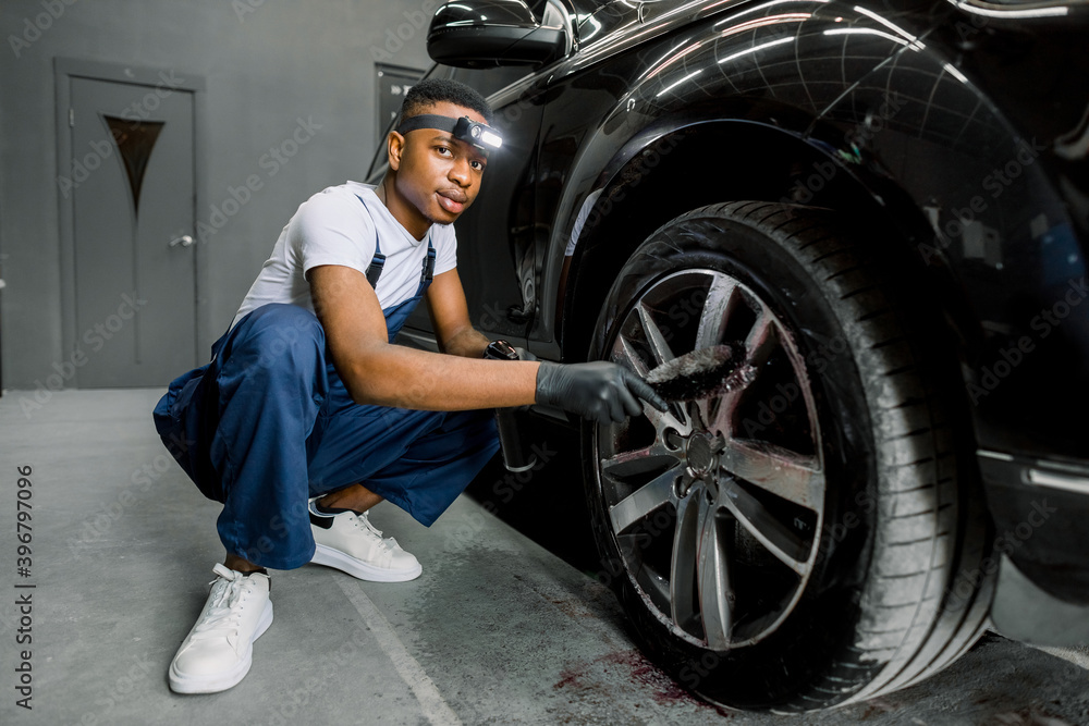Professional African male worker in overalls and black protective gloves, cleaning alloy wheels rims of luxury car with a special brush for cast wheels in a detailing workshop, looking at camera Professional African male worker in overalls and black protective gloves, cleaning alloy wheels rims of luxury car with a special brush for cast wheels in a detailing workshop, looking at camera