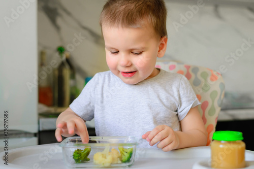 Happy 2-year-old boy kid eats cauliflower, broccoli and mashed potatoes in a jar in his kitchen at home. Baby food, solid food concept.