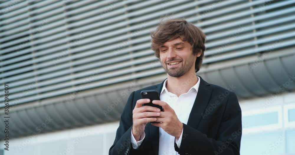 Portrait of Handsome Caucasian Successful Businessman is smiling Charmingly while browsing the Internet. Happy Caucasian Man is using Smartphone outdoor in Business District, Wearing stylish Suit. Portrait of Handsome Caucasian Successful Businessman is smiling Charmingly while browsing the Internet. Happy Caucasian Man is using Smartphone outdoor in Business District, Wearing stylish Suit.