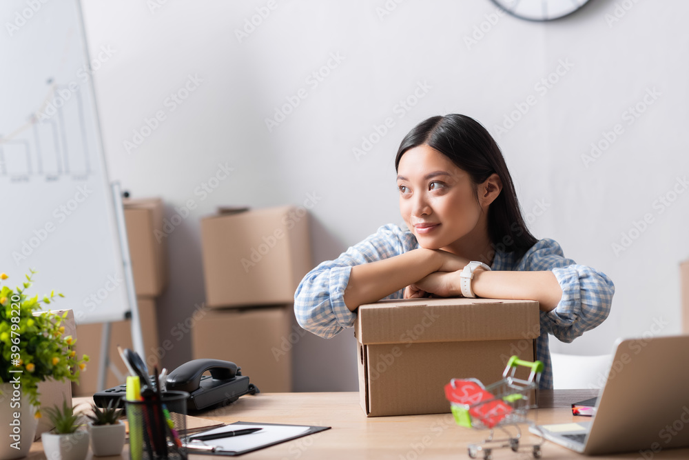 Smiling asian volunteer looking away while leaning on carton box on desk in charity center on blurred foreground Smiling asian volunteer looking away while leaning on carton box on desk in charity center on blurred foreground
